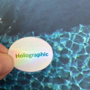 May include: A round, holographic sticker with the word "Holographic" printed in rainbow colors. The sticker has a reflective, iridescent surface. The background is a blue and teal pattern.