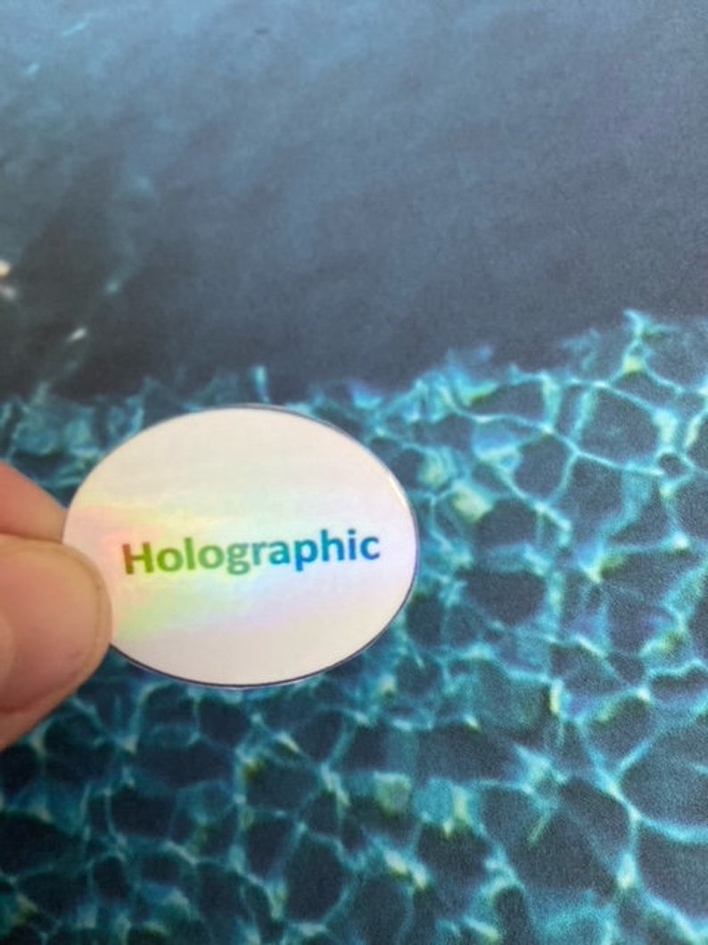 May include: A round, holographic sticker with the word "Holographic" printed in rainbow colors. The sticker has a reflective, iridescent surface. The background is a blue and teal pattern.