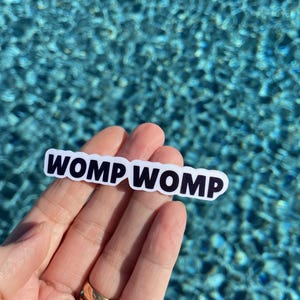 May include: A white sticker with the text "WOMP WOMP" in black lettering. The sticker is being held by a hand with a gold ring.