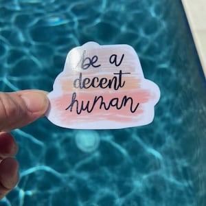 May include: A white sticker with a watercolor-style background in shades of peach, orange, and beige. The sticker features the text "be a decent human" in black cursive lettering.