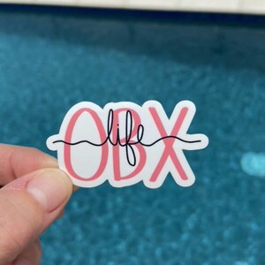 May include: A white sticker with pink lettering that says "OBX life" with a wavy line under the word "life".