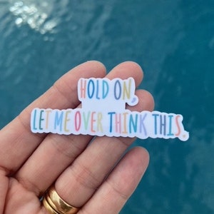 May include: A white sticker with the text "Hold on, let me overthink this" in colorful letters. The sticker is being held by a hand with a gold ring.