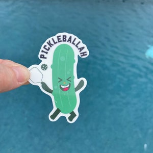 May include: A green pickle character wearing a white headband and holding a white paddle with a black ball. The pickle is smiling and has a black outline. The text "Pickleballah" is written in black around the pickle.