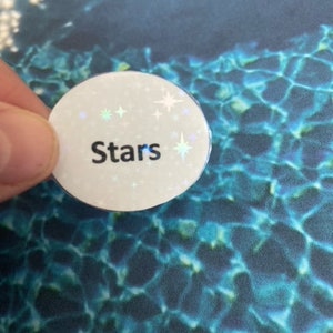 May include: A white, circular button with the word "Stars" in black, surrounded by small, iridescent star-shaped accents. The button is held against a blue and teal patterned background.