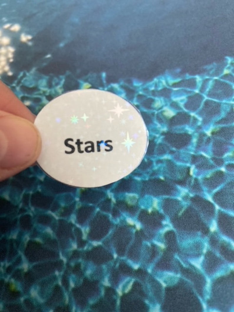 May include: A white, circular button with the word "Stars" in black, surrounded by small, iridescent star-shaped accents. The button is held against a blue and teal patterned background.