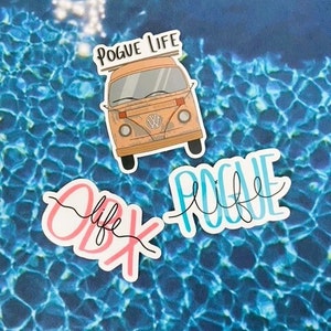 May include: Three stickers with a beach theme. The first sticker is a cartoon of a vintage van with the text "Pogue Life" above it. The second sticker is the text "OBX Life" in pink and black. The third sticker is the text "Pogue Life" in blue and black.