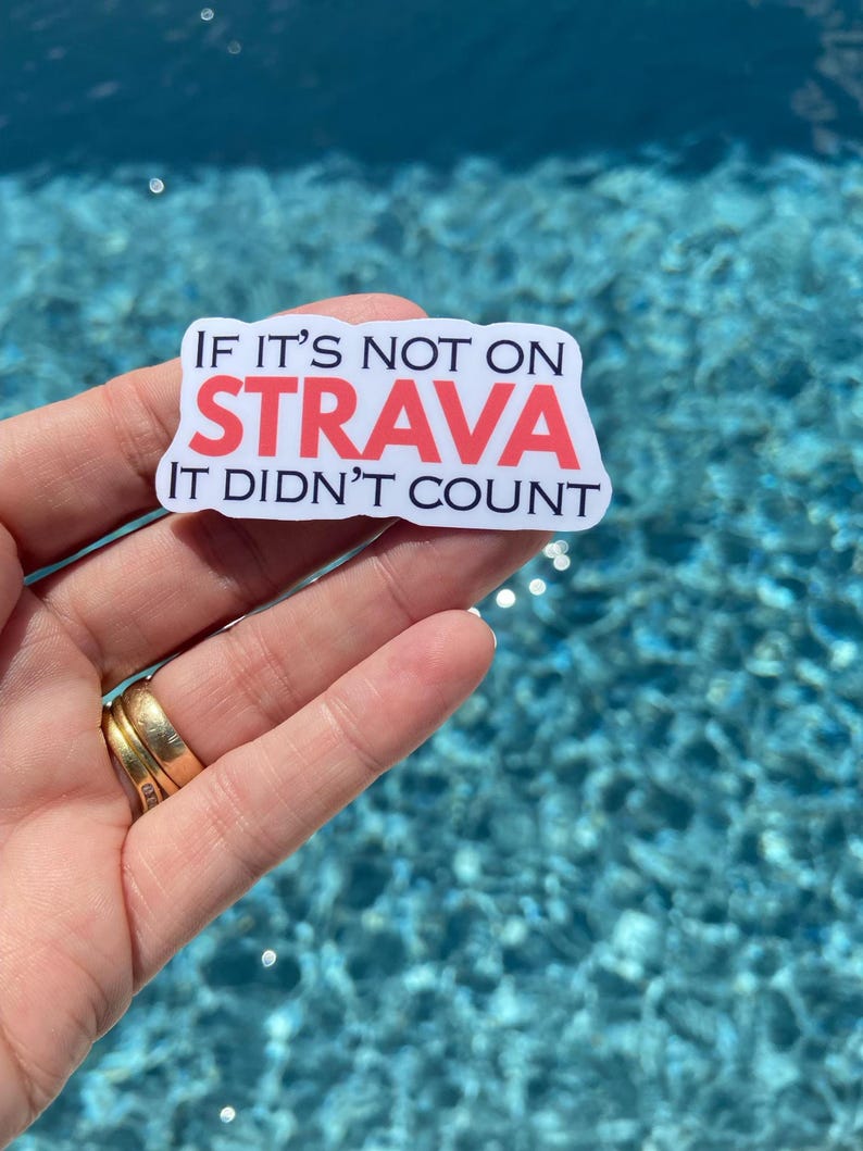 May include: A rectangular sticker with the text "IF IT'S NOT ON STRAVA IT DIDN'T COUNT" in black and red. The sticker is held in front of a blue water background.