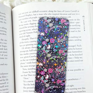 May include: A decorative bookmark with a dark background featuring bats, moons, and flowers. The bookmark is rectangular and is placed on top of an open book.