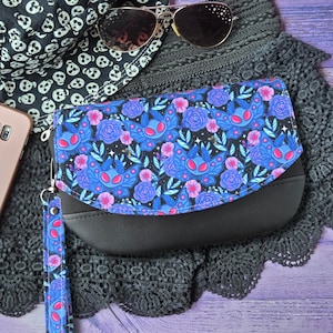 May include: A black clutch bag with a flap featuring a vibrant blue and pink moth and floral pattern, accompanied by a matching wrist strap. The bag is displayed alongside a black and white skull-patterned bucket hat and aviator sunglasses.