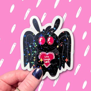 May include: A black moth sticker with iridescent glitter accents. The moth has large red eyes and is holding a red heart with the words "Live Laugh Lark". The sticker is on a pink background with white streaks.