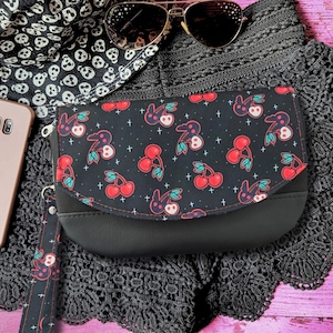May include: A black wristlet clutch featuring a flap with a repeating pattern of red cherries and white skull-shaped cherries on a black background, accented with small white crosses. The clutch includes a matching wrist strap and a solid black base.