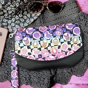 May include: A black wristlet pouch featuring a flap with a checkered pattern and circular icons of various creatures including a dinosaur, octopus, and rabbit. The pouch has a matching wrist strap and a solid black base with a smooth texture.