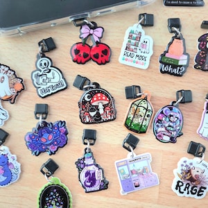 May include: A collection of colorful phone charms with various designs, including cherries, a mushroom, a ghost, and a cat on books. Some charms have text like "Read More" and "What?"