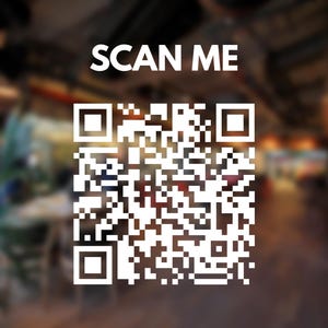 May include: White QR code with the text "SCAN ME" in a blurred indoor setting. The QR code is a square pattern of black and white squares. The background is a blurry view of a room with tables and plants.