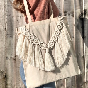 May include: A beige canvas tote bag with a large, white macrame design and fringe trim. The macrame design features a V-shape with a tassel at the bottom.