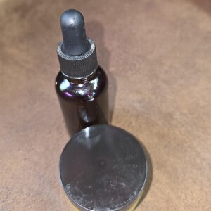 May include: A dark brown glass bottle with a black dropper and a round, dark-colored jar with a silver lid. The products are set against a brown background, suggesting a skincare or cosmetic theme.