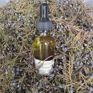 May include: A small, clear glass bottle of organic juniper berry oil with a black dropper. The bottle is surrounded by dried juniper branches and berries. The label reads "Organic Juniper Berry Oil."