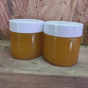 May include: Two small, clear glass jars filled with a golden-orange substance, each topped with a white lid. The jars sit on a wooden surface against a backdrop of textured wood. The contents of the jars are visible, suggesting a product like honey or a cosmetic.