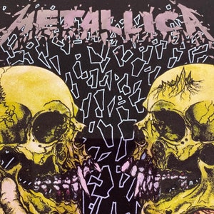 K&ouml;nnte beinhalten: Schwarzes T-Shirt mit einem Motiv aus zwei gelben Sch&auml;deln mit Z&auml;hnen und einer rosa Zunge. Das Wort "METALLICA" ist in einem gebrochenen, fragmentierten Stil in Lila und Wei&szlig; geschrieben.