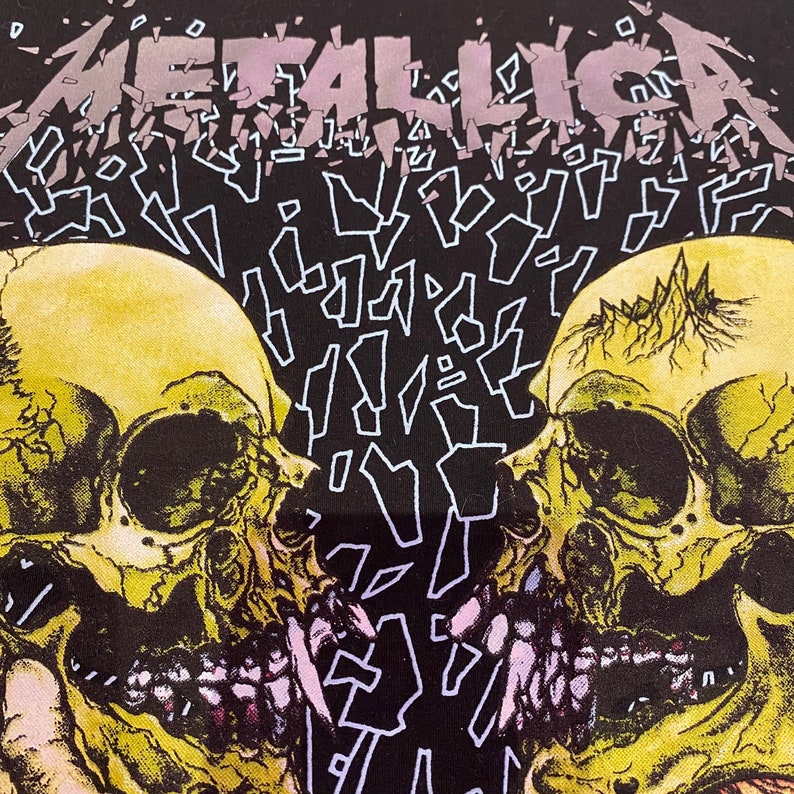 K&ouml;nnte beinhalten: Schwarzes T-Shirt mit einem Motiv aus zwei gelben Sch&auml;deln mit Z&auml;hnen und einer rosa Zunge. Das Wort "METALLICA" ist in einem gebrochenen, fragmentierten Stil in Lila und Wei&szlig; geschrieben.