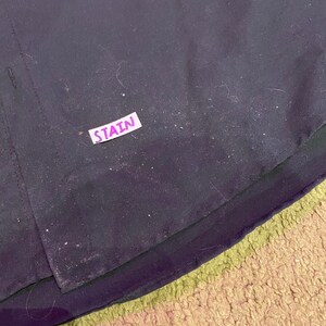 K&ouml;nnte beinhalten: Nahaufnahme einer schwarzen Stoffoberfl&auml;che mit einem wei&szlig;en Etikett, auf dem "STAIN" in lila Buchstaben steht.