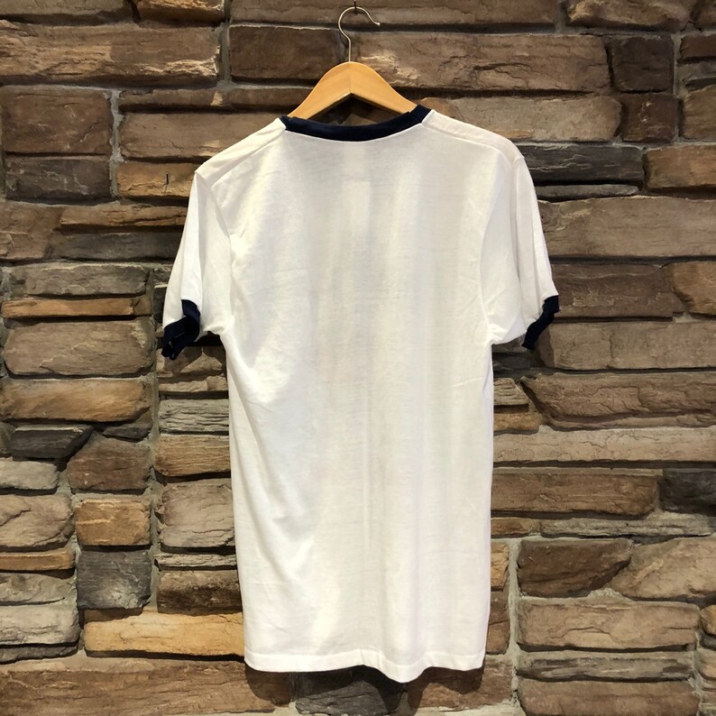 May include: White t-shirt with a navy blue ringer collar and cuffs. The shirt is hanging on a wooden hanger against a brick wall.