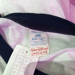 May include: A white and pink striped t-shirt with a navy blue ribbed collar and cuffs. The shirt has a white tag with the text "TROPIX TOGS" and "WALT DISNEY CHARACTER" printed on it. The tag also includes the text "SHRINKAGE CONTROLLED 50% POLYESTER 50% COTTON" and "LARGE MADE IN USA".