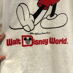 May include: White t-shirt with a black and white cartoon image of Mickey Mouse. The shirt has red text that says "Walt Disney World" and a red Mickey Mouse head with a globe inside.