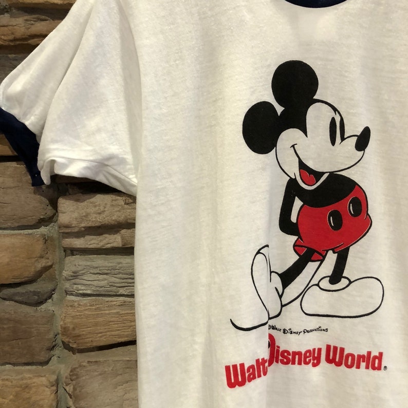 May include: White t-shirt with a blue ringer neck. The shirt features a black and white illustration of Mickey Mouse wearing red shorts. The text "Walt Disney World" is printed in red below the illustration.