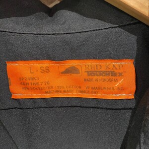 K&ouml;nnte beinhalten: Ein orangefarbenes und schwarzes Kleidungsetikett mit dem Text "RED KAP" und "MADE IN HONDURAS". Das Etikett enth&auml;lt auch den Text "L-SS", "SP24BK3", "4CM 1H6776", "98% POLYESTER/35% COTTON", "VF IMAGEWEAR, INC" und "MACHINE WASH TUMBLE DRY".