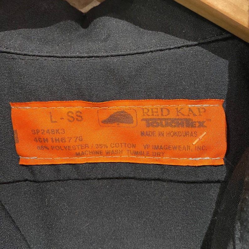 K&ouml;nnte beinhalten: Ein orangefarbenes und schwarzes Kleidungsetikett mit dem Text "RED KAP" und "MADE IN HONDURAS". Das Etikett enth&auml;lt auch den Text "L-SS", "SP24BK3", "4CM 1H6776", "98% POLYESTER/35% COTTON", "VF IMAGEWEAR, INC" und "MACHINE WASH TUMBLE DRY".