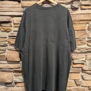 May include: A dark grey t-shirt hangs on a wooden hanger against a brick wall. The shirt is oversized with short sleeves and a crew neck. The fabric appears soft and slightly worn.