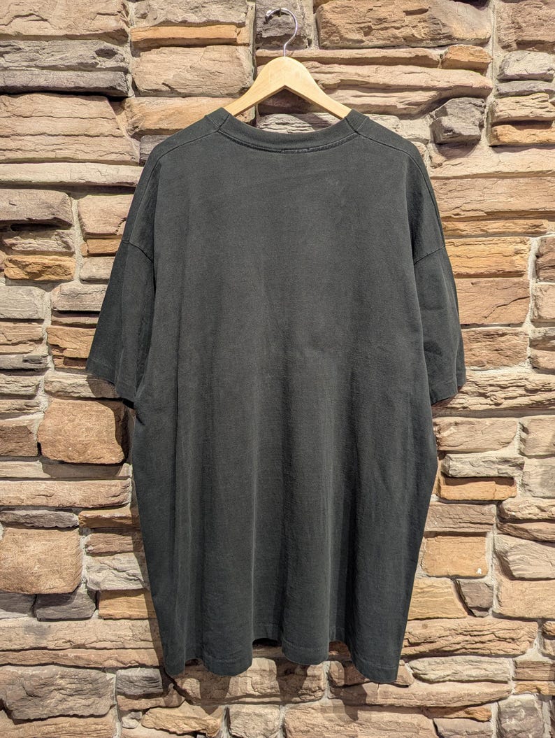 May include: A dark grey t-shirt hangs on a wooden hanger against a brick wall. The shirt is oversized with short sleeves and a crew neck. The fabric appears soft and slightly worn.