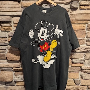 May include: Black t-shirt with a Mickey Mouse graphic. The cartoon character is shown in a dynamic pose, wearing white gloves, a red shirt, and yellow shoes. The t-shirt is displayed on a wooden hanger against a brick wall background.