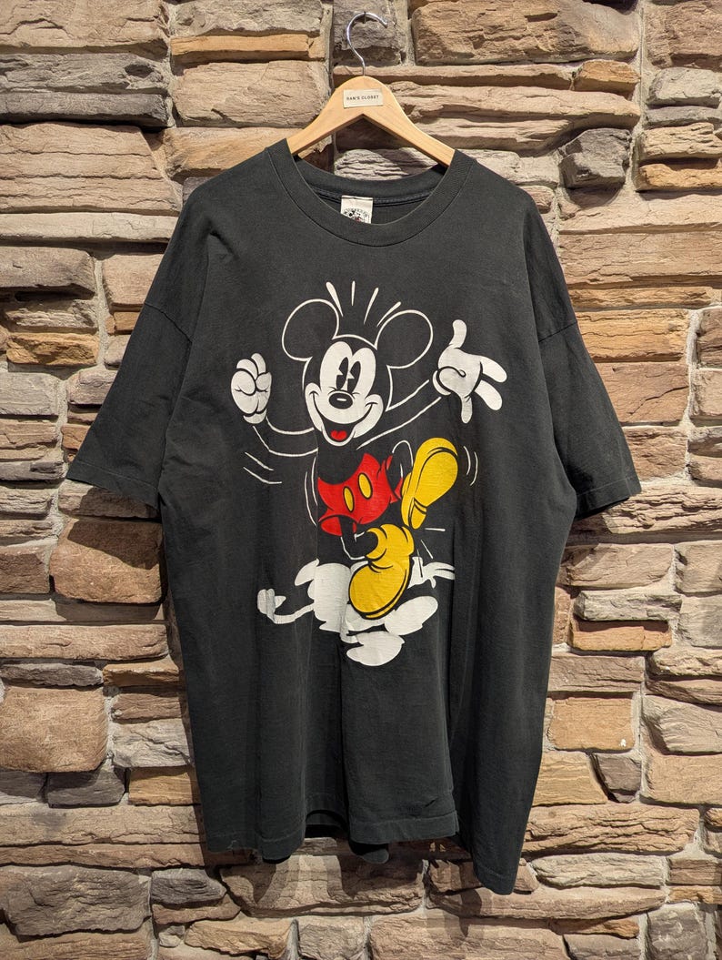 May include: Black t-shirt with a Mickey Mouse graphic. The cartoon character is shown in a dynamic pose, wearing white gloves, a red shirt, and yellow shoes. The t-shirt is displayed on a wooden hanger against a brick wall background.
