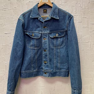 May include: A vintage blue denim jacket with a classic design. The jacket has two chest pockets, button fastenings, and a collar. The Lee brand label is visible. The jacket is hanging on a wooden hanger.