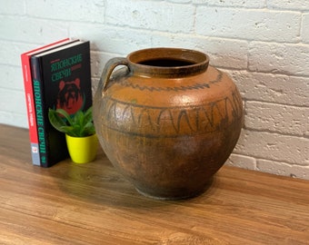 May include: A large, brown, earthenware pot with a wide mouth and two handles. The pot has a decorative pattern of brown lines and swirls around the top and bottom. The pot is sitting on a wooden table.