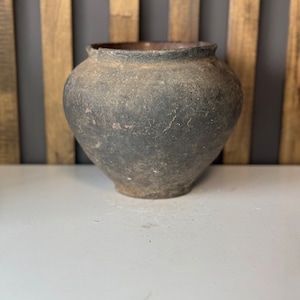 Op de afbeelding: Een ronde, donkerbruine, keramische pot met een ruw, getextureerd oppervlak. De pot staat op een wit oppervlak.