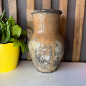 Könnte beinhalten: Eine rustikale braune und beigefarbene Keramikvase mit einer abgenutzten, strukturierten Oberfläche. Die Vase hat einen breiten Boden und einen schmalen Hals.