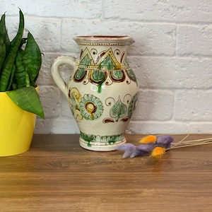 May include: A white ceramic pitcher with a brown rim and green and brown floral designs. The pitcher has a handle and a rounded body.