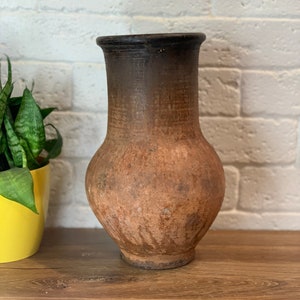 May include: A brown clay vase with a dark brown rim. The vase has a rounded body and a narrow neck. It is sitting on a wooden surface.