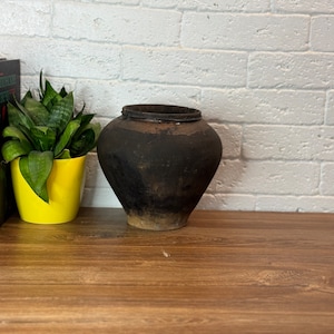 May include: A dark brown clay pot with a wide mouth sits on a wooden table in front of a white brick wall. A potted green plant and two books are on the table to the left of the pot.