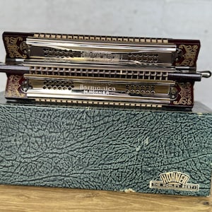 Könnte beinhalten: Eine Vintage-Hohner-Mundharmonika in dunkelroter Ausführung mit goldenen Akzenten, die auf einer strukturierten grünen Schachtel liegt. Die Mundharmonika hat eine silberne Metallplatte mit dem Text "Harmonica M. Hohner". Die Schachtel trägt den Text "Hohner The World's Best".