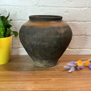 May include: A large, dark brown, earthenware pot with a rounded body and a wide opening. The pot has a rustic, aged appearance and is sitting on a wooden surface.