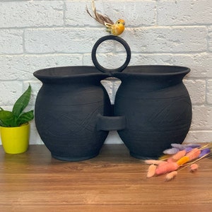 Puede incluir: Dos macetas de cerámica negra con un diseño decorativo, conectadas por un anillo de metal. Una pequeña figurita de pájaro amarillo se sienta en la parte superior del anillo.