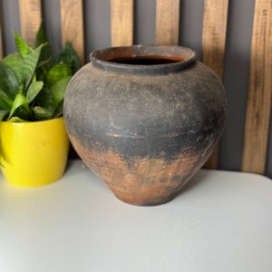 May include: A large, dark brown, earthenware pot with a rounded body and a wide opening. The pot has a rustic, aged appearance and is sitting on a white surface.