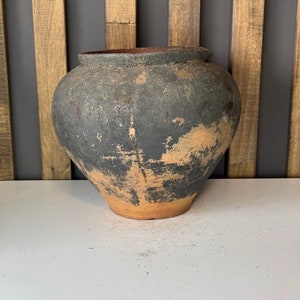 May include: A large, dark brown, earthenware pot with a wide mouth and a rounded body. The pot has a rustic, aged appearance with a mottled surface.