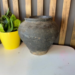 May include: A dark brown, round, ceramic pot with a slightly flared rim. The pot has a rustic, aged appearance with a textured surface.