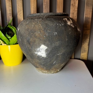 Puede incluir: Una maceta grande, redonda y de cerámica de color marrón oscuro con una superficie rugosa y texturizada. La maceta tiene manchas blancas en la superficie y está sentada sobre una superficie blanca.