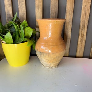 Peut inclure: Un vase en terre cuite avec un glaçage brun clair. Le vase a un corps arrondi et un col étroit. Il est posé sur une surface blanche.